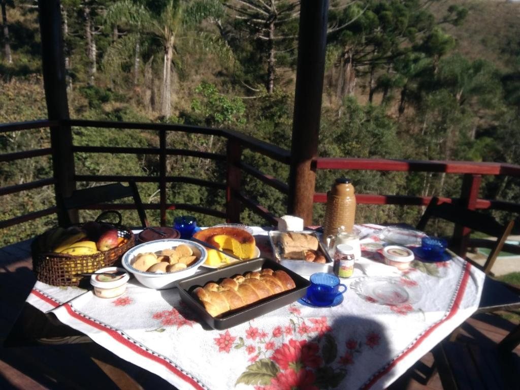 Mesa de café da manhã na varanda, com pães, bolo e frutas, de frente para a mata de araucárias